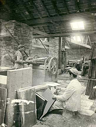 Photograph: Slate-making: splitter is parting a block with special slate-splitting chisel. His partner dresses the slates to size on the revolving knife machine.